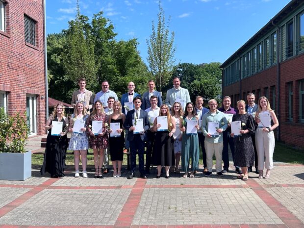 Eine Gruppe von Menschen steht im Freien in zwei Reihen, hält Urkunden in den Händen und lächelt in die Kamera. Die Kulisse ist sonnig, im Hintergrund sind Backsteingebäude, Bäume und ein blauer Himmel zu sehen.